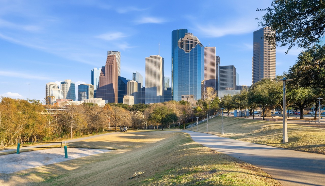 Panoramic view of the downtown Houston skyline over a green park and walking paths under a clear blue sky
