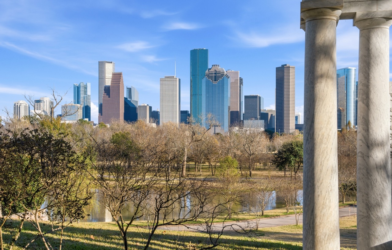 View of the Houston downtown skyline across a serene park pond with mature trees under bright sun